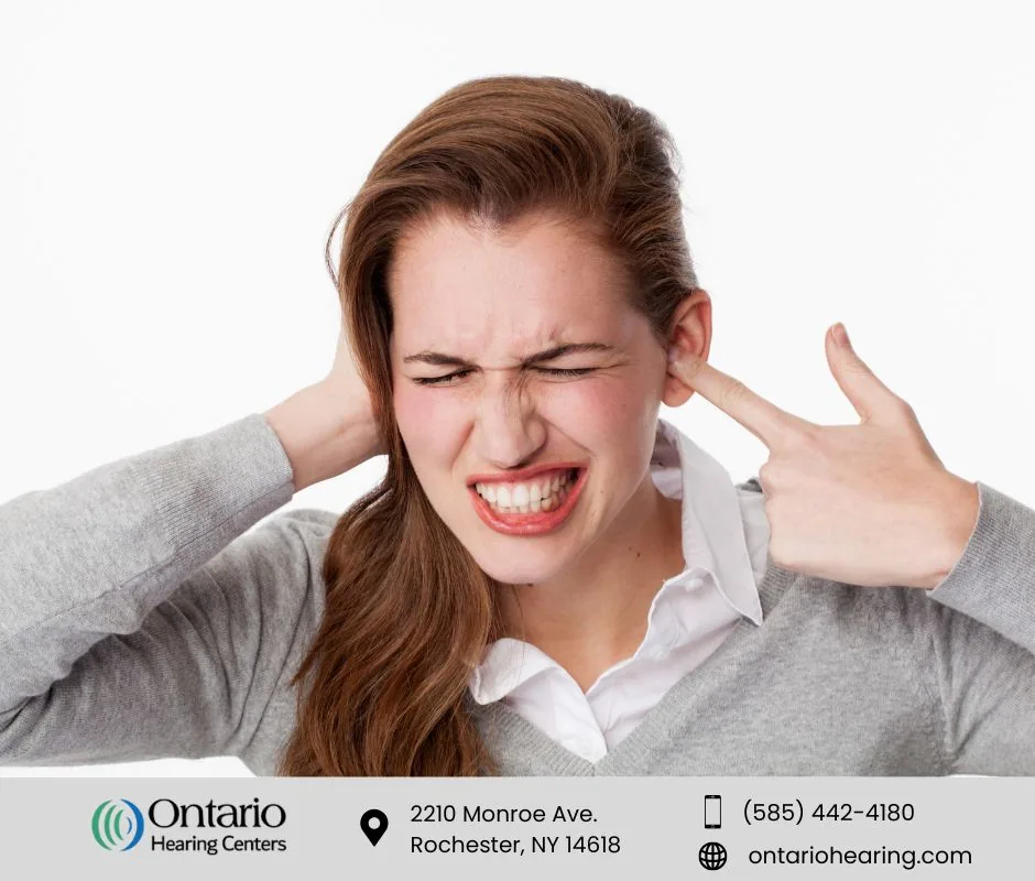 Woman grimacing in pain while covering one ear and pointing to the other, indicating ear discomfort or noise sensitivity.